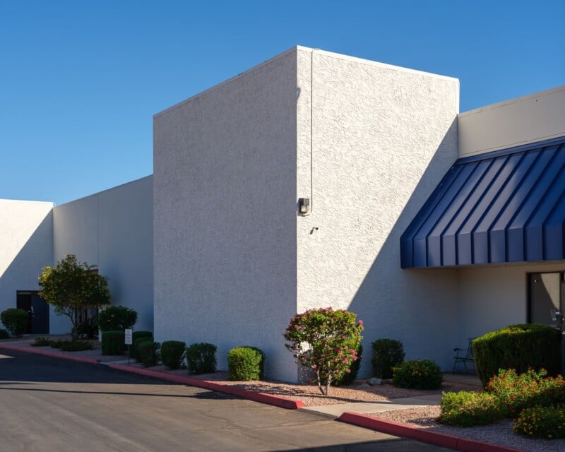 A crisp image of a modern commercial building corner with textured white walls under a clear blue sky, featuring shrubs and blooming flowers along the sidewalk.