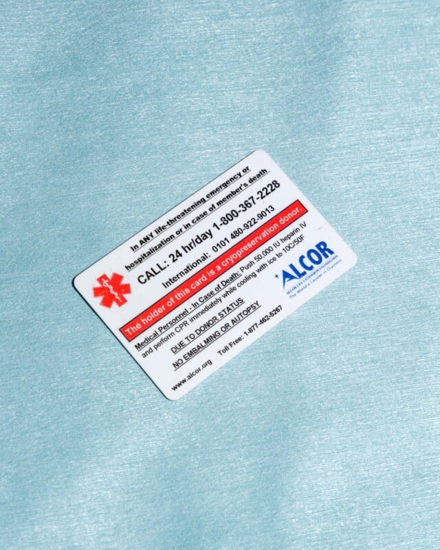 An alcor membership card on a textured blue background, displaying emergency contact numbers and a red star logo. text includes details for 24-hour medical support.