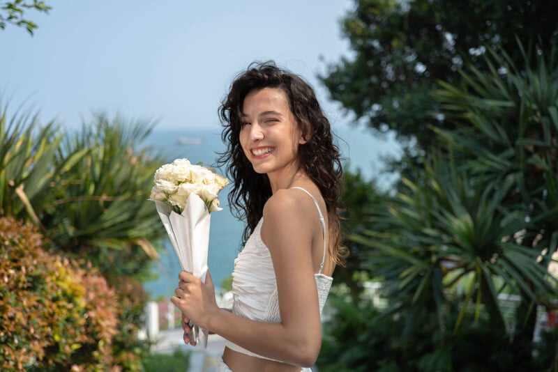 A joyful bride holding a bouquet of white flowers, smiling outdoors with lush greenery and the ocean in the background.