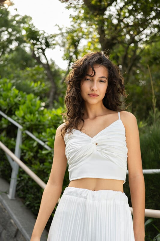 A young woman with curly hair stands outdoors, wearing a white top and skirt, with a backdrop of lush greenery and a staircase.