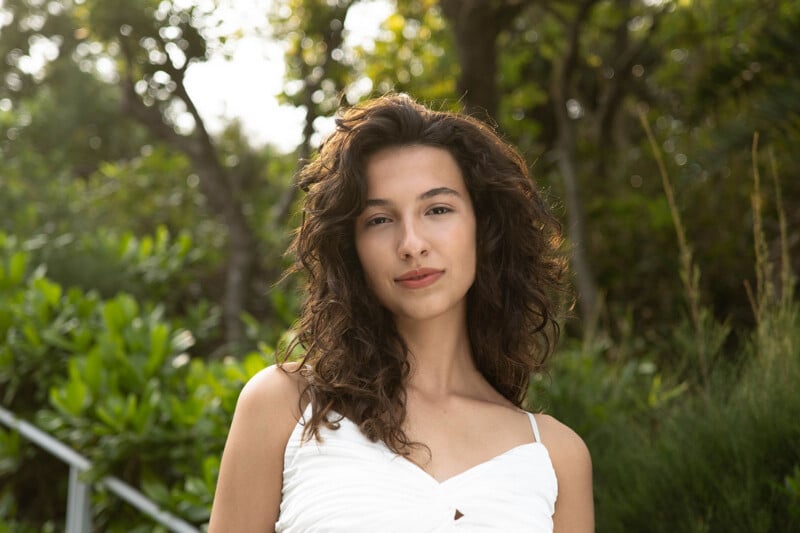 A young woman with curly brown hair and a white dress stands outdoors, surrounded by lush greenery, looking directly at the camera with a soft expression.