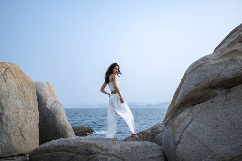 A woman in a white dress stands between large rocks on a beach, with the ocean and a hazy sky in the background. she looks away from the camera, her hair tousled by the wind.