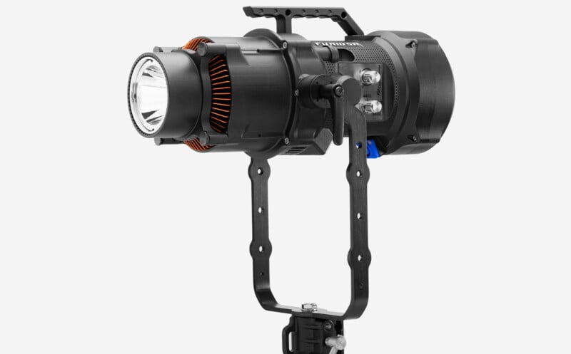 A high-resolution photo of a modern, professional-grade spotlight with a detailed design, featuring a black body, prominent lens, and orange accents, mounted on an adjustable black handle.