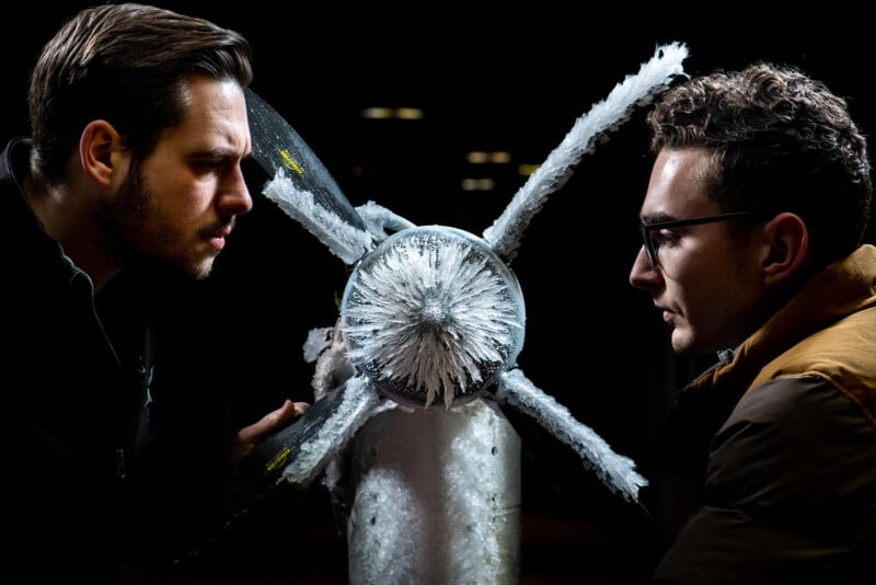 Two men intensely facing each other with a shattered glass object between them, highlighted by dramatic lighting in a dark setting.