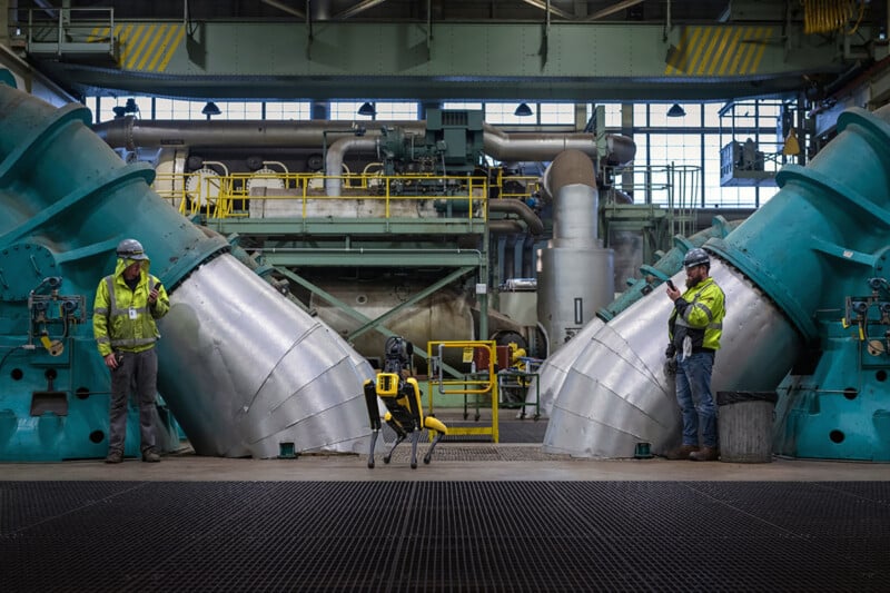 Two workers inspect large metallic ducts at an industrial plant. they wear high-visibility vests and hard hats, with tools and equipment scattered around the workspace.