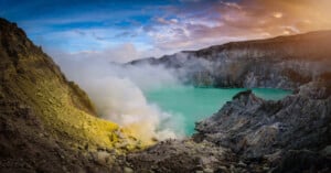 Panoramic view of a stunning turquoise crater lake with rising steam, surrounded by rugged cliffs under a dramatic sky at sunrise.