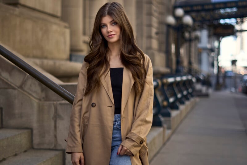 A young woman in a stylish trench coat smiling, standing on a city street with buildings and an iron railing in the background.