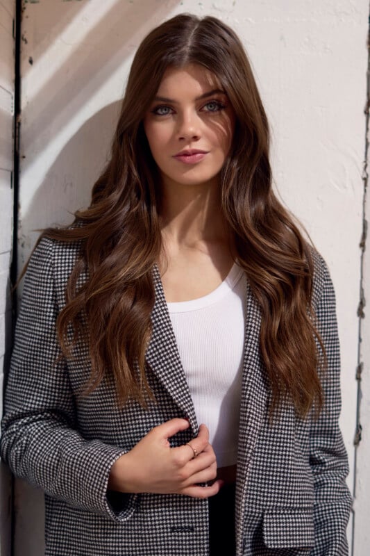A young woman with long, wavy brown hair, wearing a houndstooth jacket and a white top, stands against a white wall, gazing intently at the camera.
