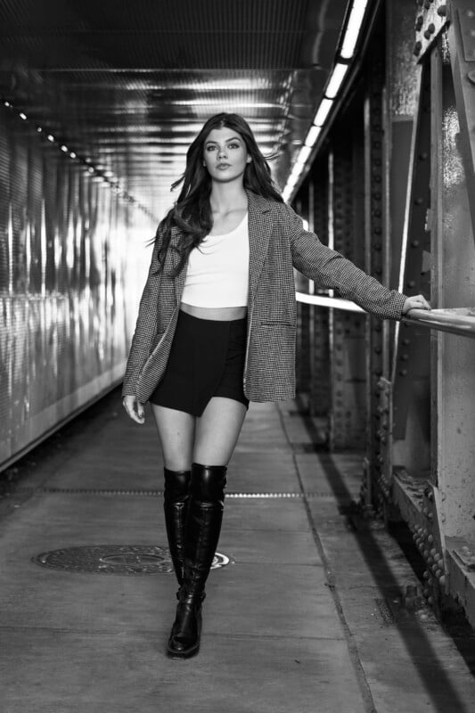 A monochrome image of a woman walking confidently down a tunnel-like corridor. she's dressed in a stylish outfit with knee-high boots, holding onto the railings.