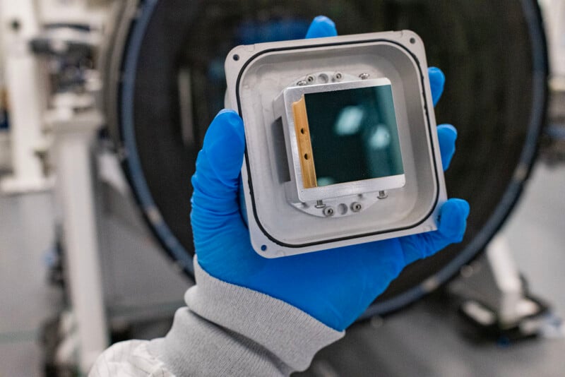 A person wearing blue gloves holding a small square device that holds a camera sensor, in a cleanroom environment with technical equipment in the background.