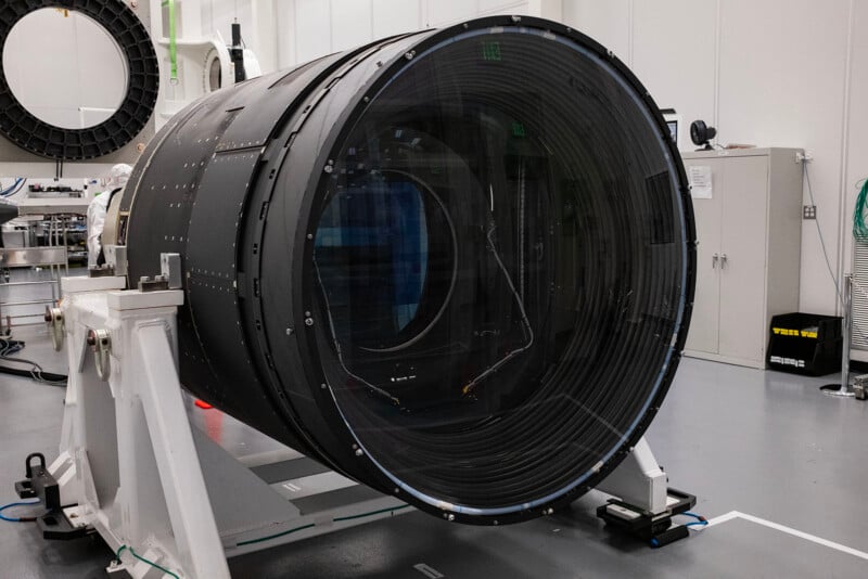 Large cylindrical LSST camera with transparent circles on both ends is positioned in a clean, modern laboratory setting, surrounded by various technical equipment.