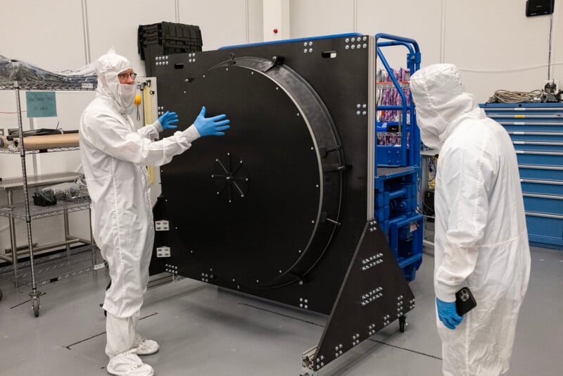 Two men in cleanroom suits inspecting a large circular black lens cap in a laboratory setting.