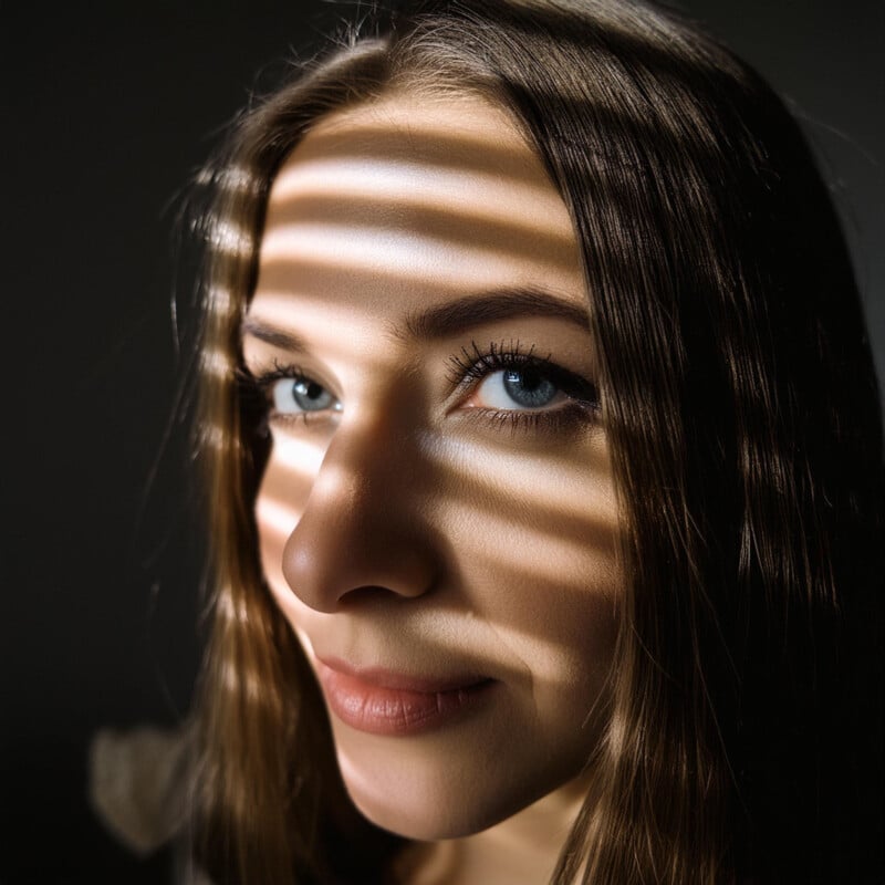 Portrait of a young woman with light skin and blue eyes, featuring shadows of blinds cast across her face. she is looking towards the light source, creating a dramatic and thoughtful expression.