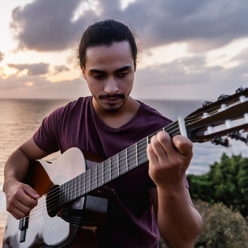 A focused young man playing an acoustic guitar outdoors at sunset, with the ocean partially visible in the background. he is wearing a purple shirt and has a thoughtful expression.
