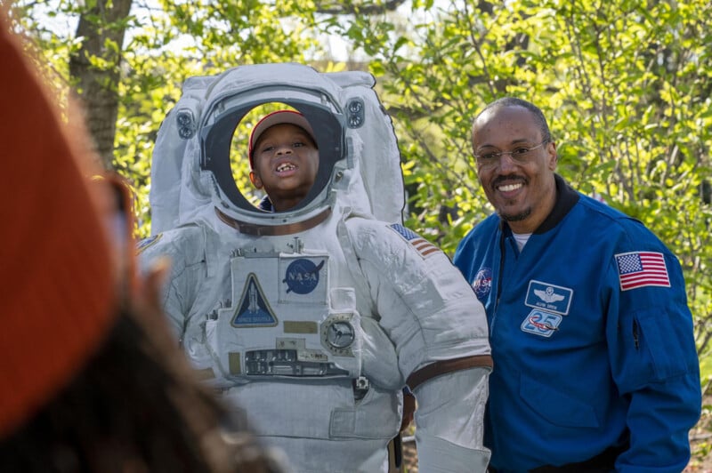 A young child in an astronaut suit stands beside an adult man in a blue nasa jumpsuit, both smiling, in a park with trees in the background.