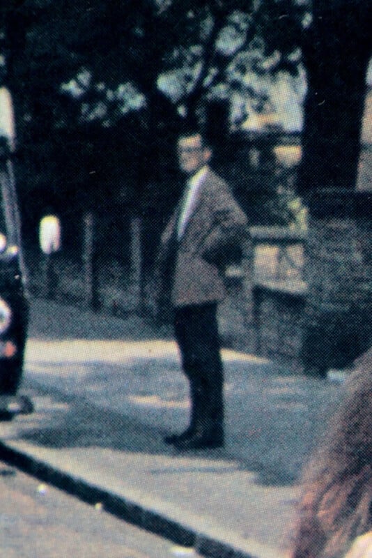 Grainy image of a man standing alone on a city sidewalk, wearing a suit and looking to the side, with blurred cars and trees in the background.