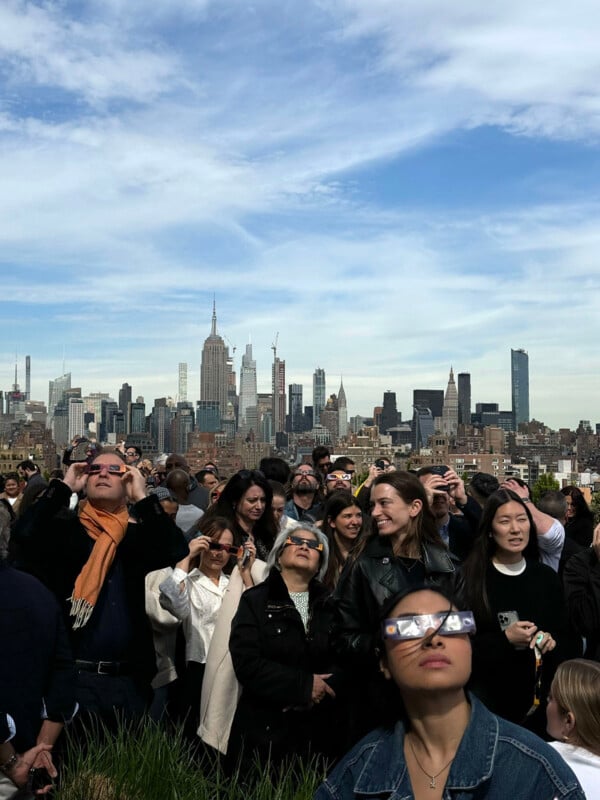 A crowd of people look up at the eclipse with the New York city skyline in the back.
