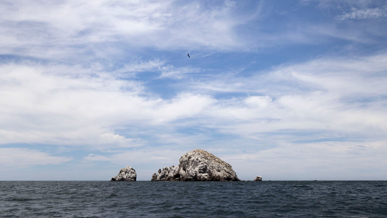 A large rugged rock formation surrounded by a slightly choppy sea under a cloudy sky, with a bird flying high above.