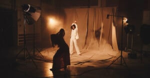 A photographer captures an energetic woman in a white suit posing on a studio set with ambient lighting and draped backdrops.