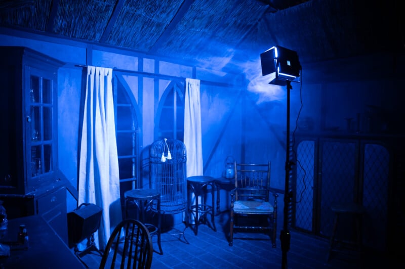 A dimly lit, blue-toned room with a thatched roof, featuring a birdcage, wooden furniture, and a spotlight casting shadows, giving a mysterious, cinematic feel.