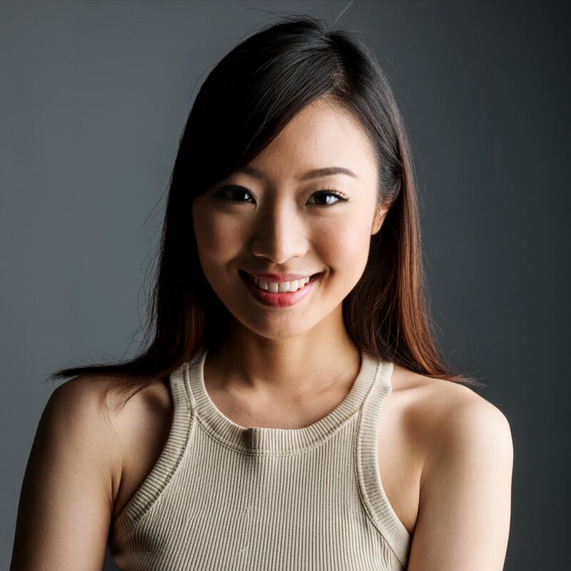 Portrait of a smiling asian woman with long hair, wearing a sleeveless beige top, against a grey background.