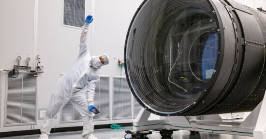 Chris Niccolls next to the LSST to demonstrate its size, gesturing upwards in a dynamic pose. the room is equipped with technical panels and vents.