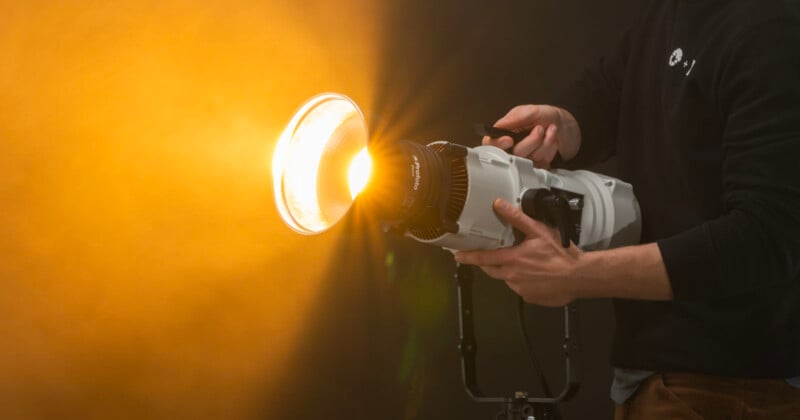 A person holding a camera with a large attached lens, aiming a bright light towards the left, in a studio with a warm, orange-hued backdrop.