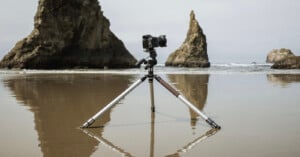 A camera mounted on a tripod positioned in shallow water on a beach, with large rock formations in the background under a cloudy sky.
