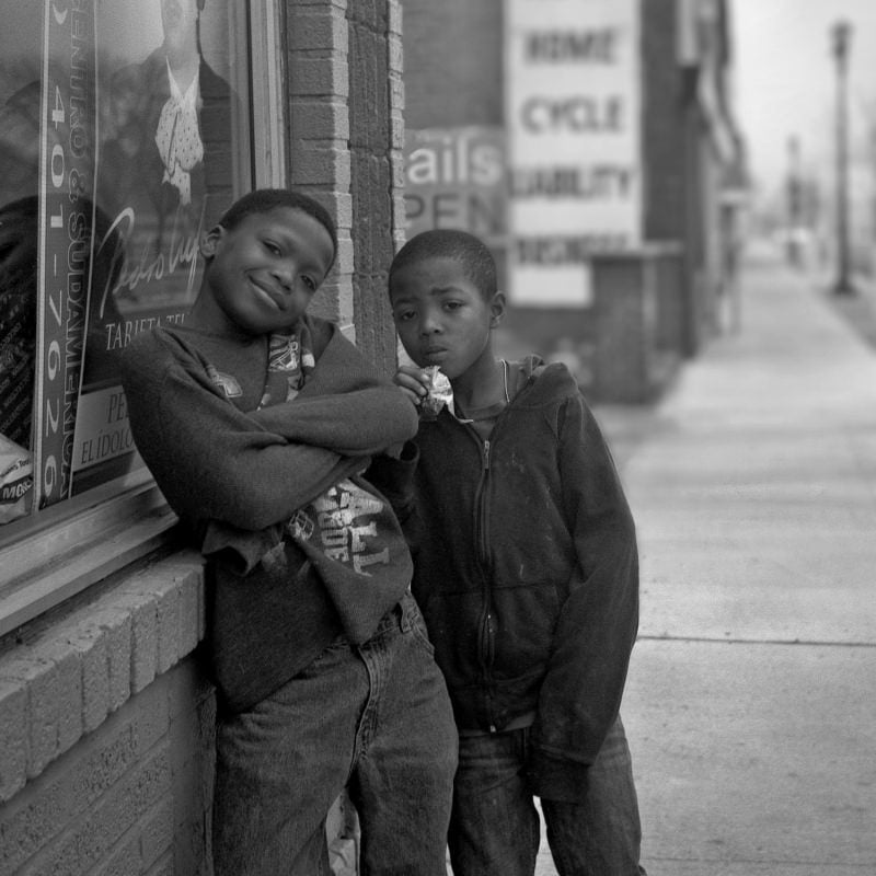Two young boys leaning against a brick wall on a city street, one smiling with his arm crossed, the other with a neutral expression, looking directly at the camera. black and white photo.