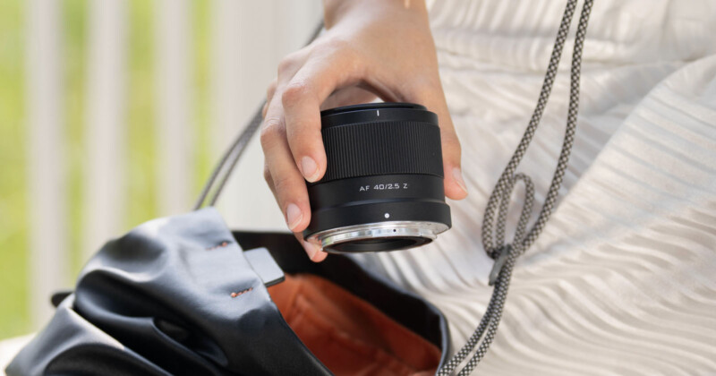 Close-up of a person's hand holding a camera lens over an open black bag, with a blurred green and white background.
