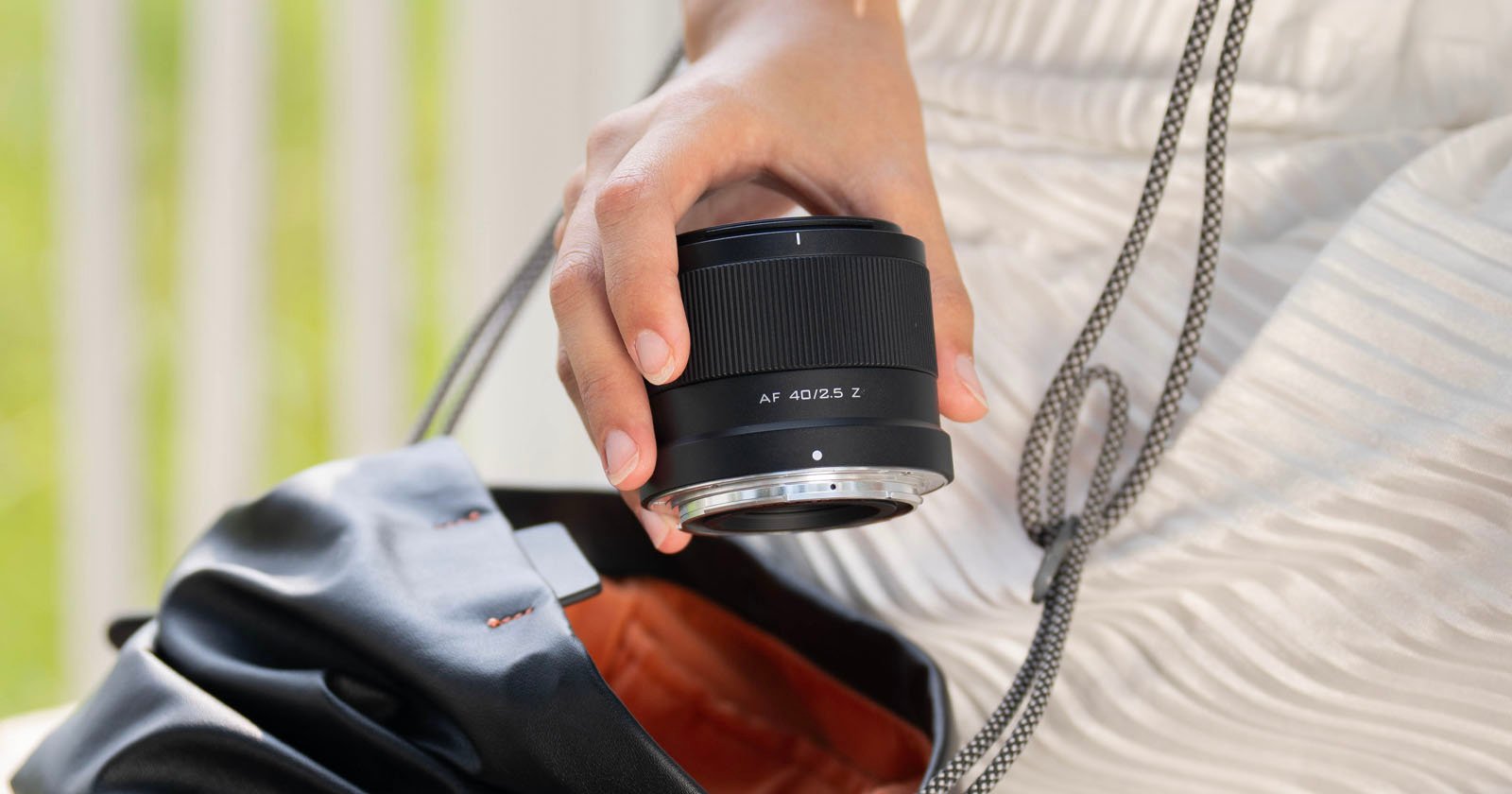 Close-up of a person's hand holding a camera lens over an open black bag, with a blurred green and white background.