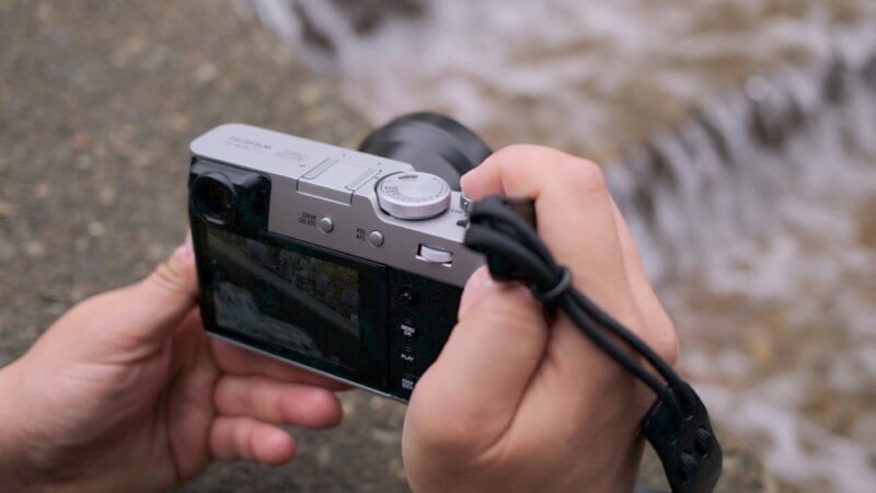 A close-up photo of a person's hands holding a digital camera, with focus on adjusting settings while capturing a stream in the background. the camera is silver with visible dials and buttons.