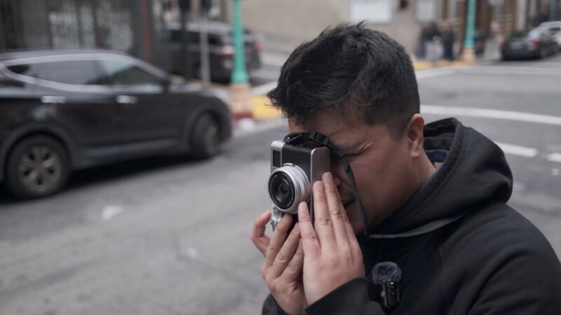 A man in a black jacket takes a photo with a vintage camera on a busy city street, focusing intently through the viewfinder.