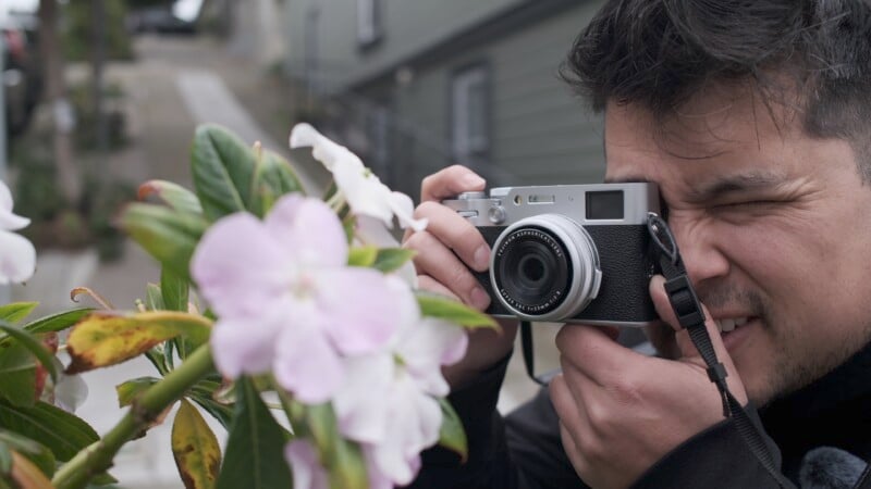 A man intently focuses on photographing pink flowers with a vintage camera, slightly obscured by the branch of blooms.