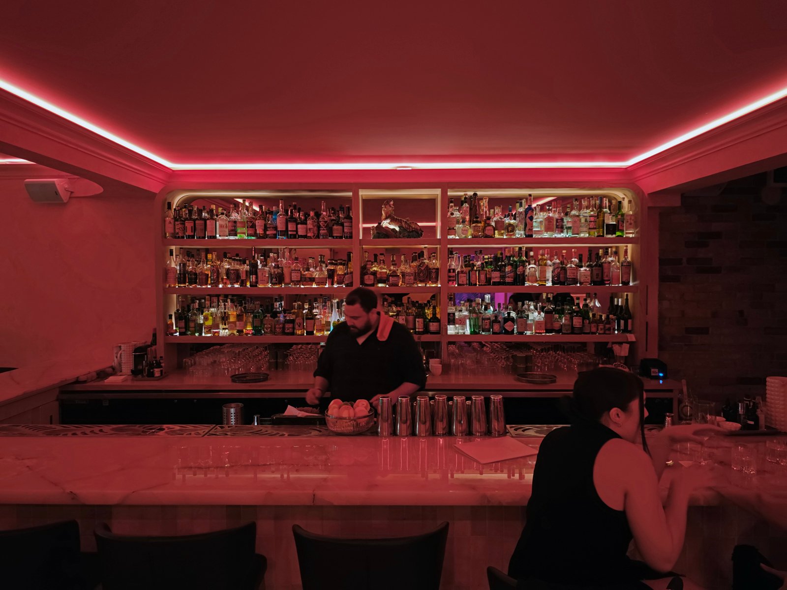 A dimly lit bar with ambient red lighting, featuring a bartender behind the bar and a customer seated at the counter. shelves stocked with various bottles line the back wall.