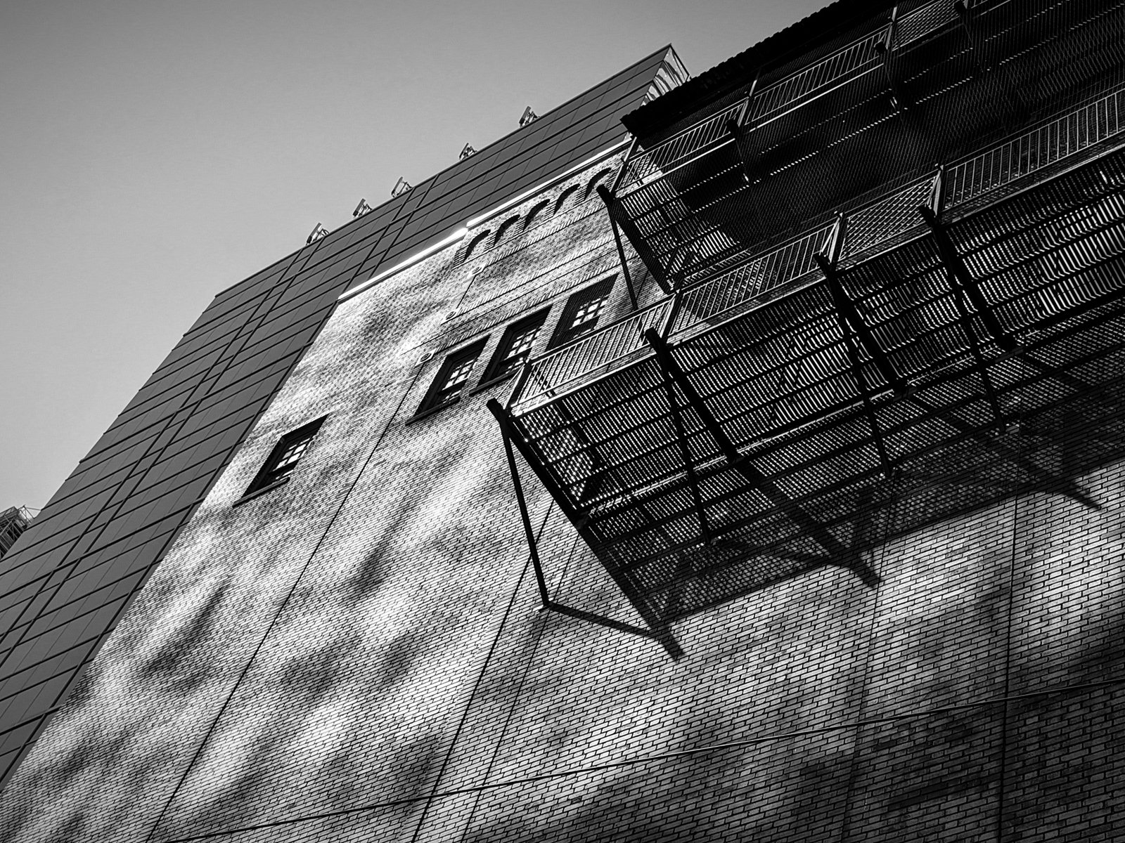 Black and white photo of a tall building with a textured facade, featuring multiple fire escapes and small windows, under a clear sky.
