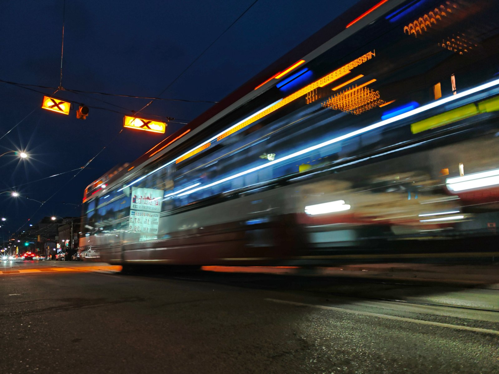 A blurred image of a streetcar in motion at night, captured from a low angle with vivid streaks of light and illuminated traffic signals overhead.