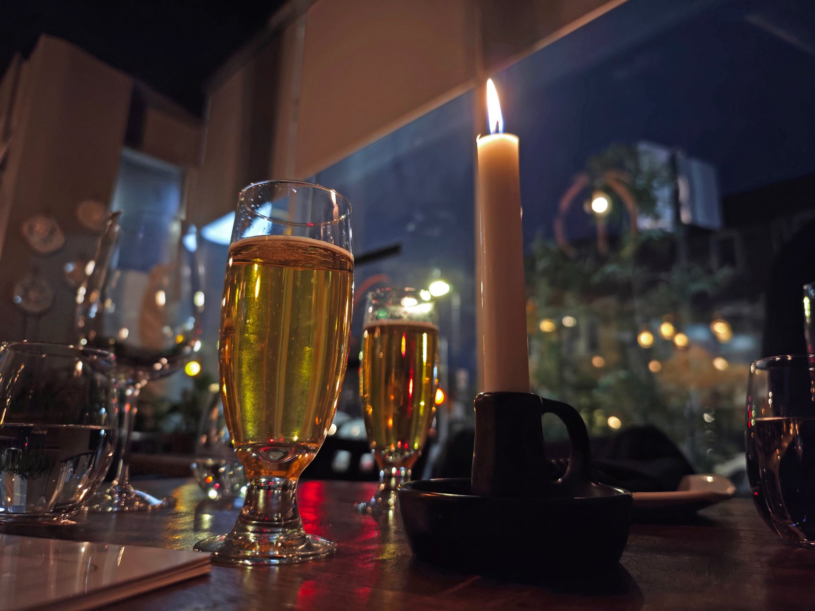 A cozy dining scene with a lit candle and two glasses of champagne on a table, with soft, blurred lights visible through a window in the background.