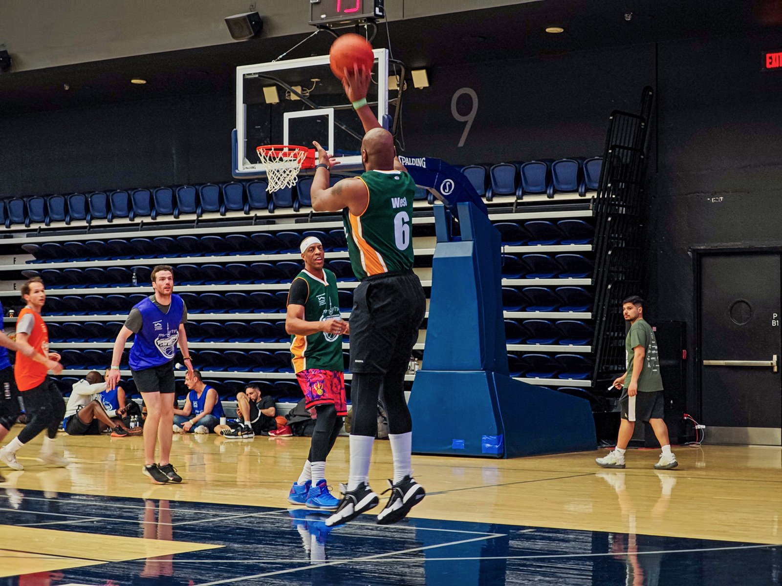 A basketball player in a green and white jersey, number 6, shooting a basketball towards the hoop during a game in an indoor court with spectators. other players are watching and waiting in various colorful jerseys.