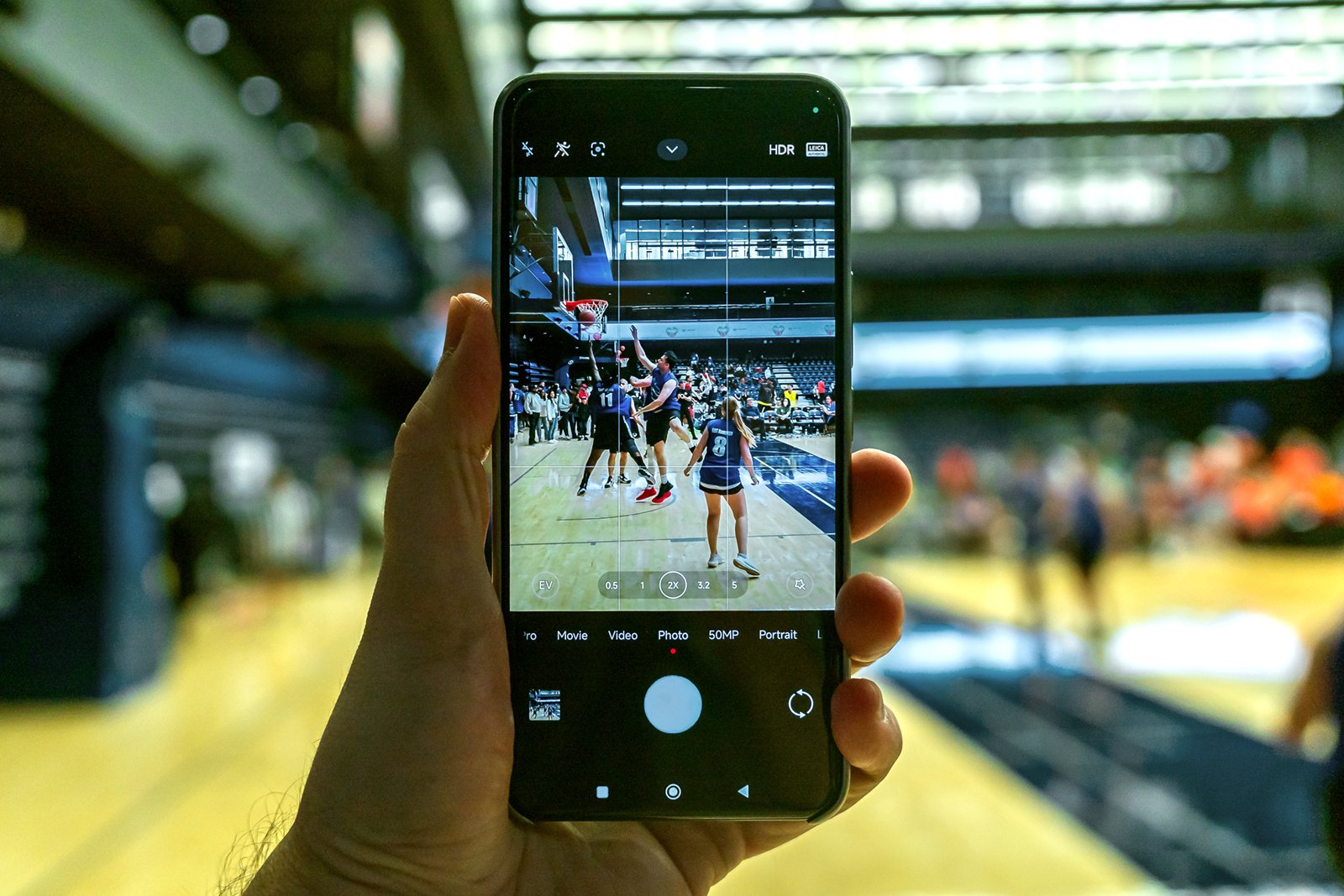 A hand holding a smartphone capturing a live basketball game, displayed on the screen with players in action and cheerleaders on the court.