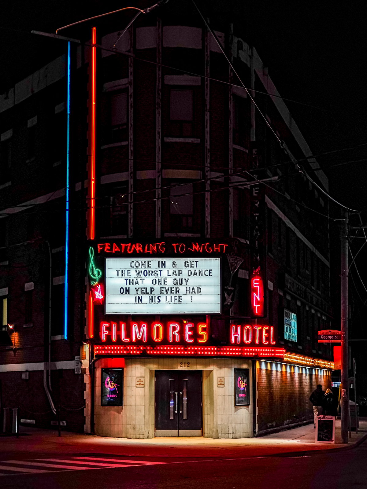 A vibrant neon sign lights up the facade of fillmores hotel at night, advertising a comedy event featuring "the worst lap dance ever" with "that one guy no one ever had on his lap.
