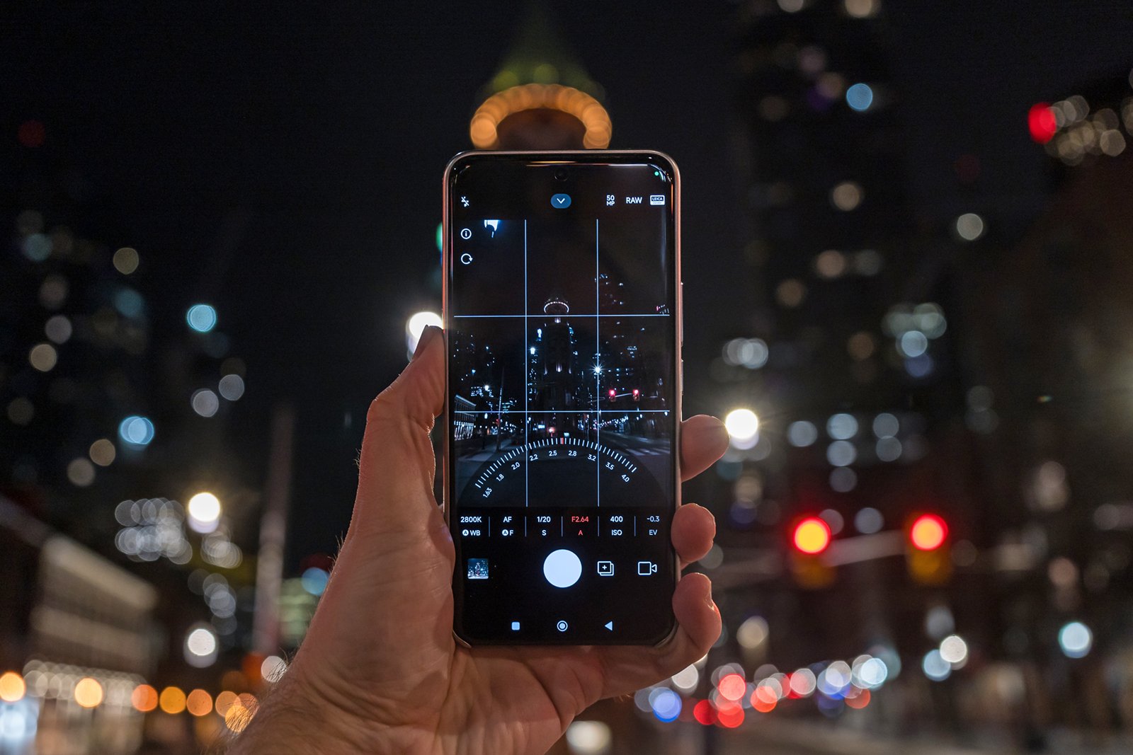 A hand holds a smartphone with its camera app open, capturing a night scene of a city with illuminated buildings and blurred lights in the background.