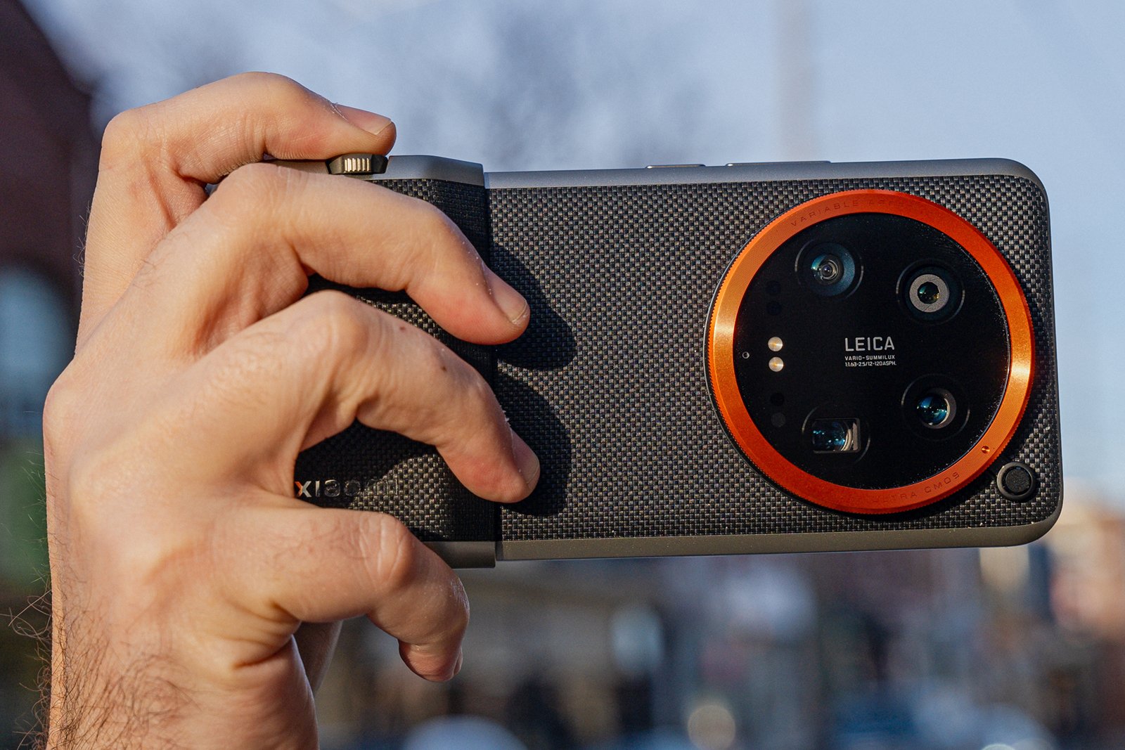 A hand holding a smartphone with a large black and red camera module labeled "leica" on the back, displayed against an outdoor blurred background.