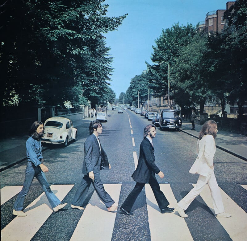Four men cross a zebra crossing in a single file on a sunny street, dressed in suits and casual wear, evoking an iconic pop culture moment. vintage cars and trees line the street.