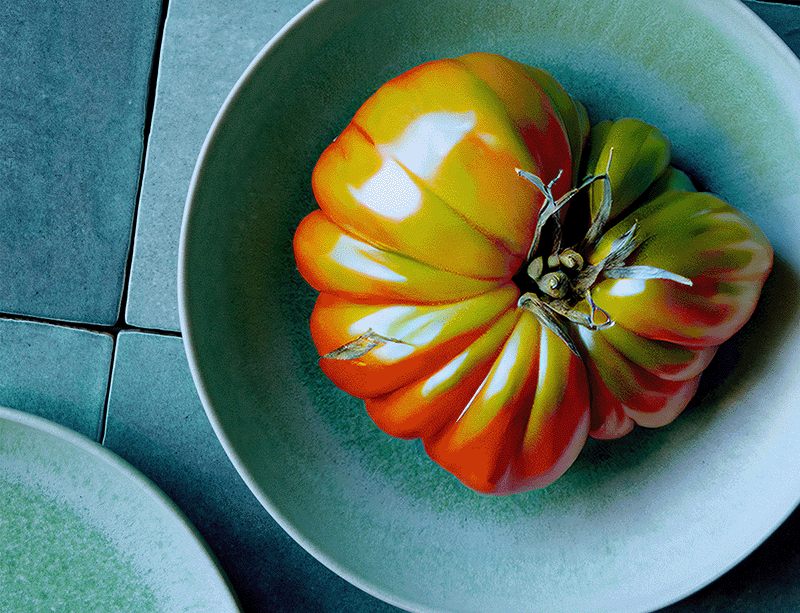 A vibrant, ribbed heirloom tomato with orange and red stripes rests in a pale green ceramic bowl on a textured blue surface, accompanied by a hint of another green plate at the edge.
