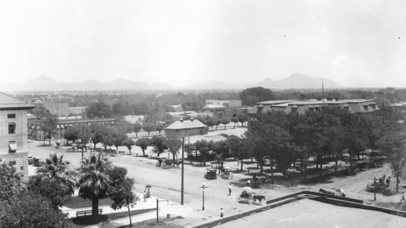 Historic black and white photo depicting an aerial view of a town square with vintage cars and sparse trees, surrounded by buildings, with mountains in the distance.