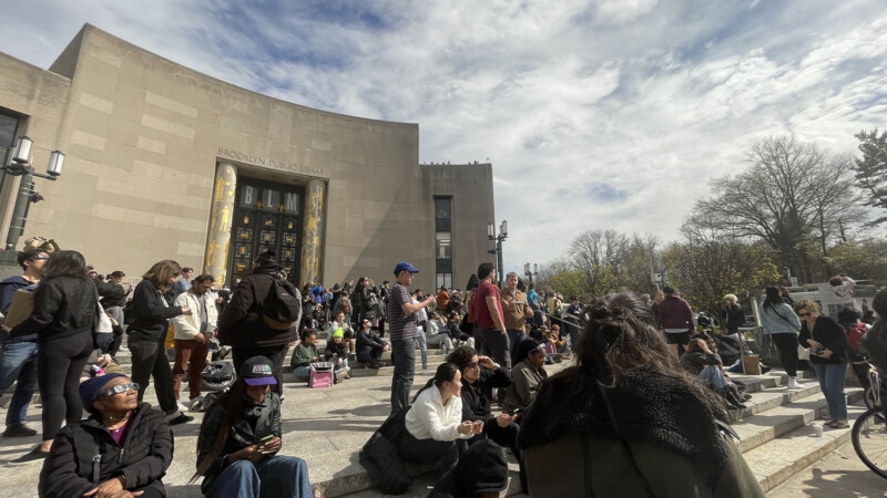 A crowd of people stand in front of the Brooklyn Public Library to watch the eclipse.