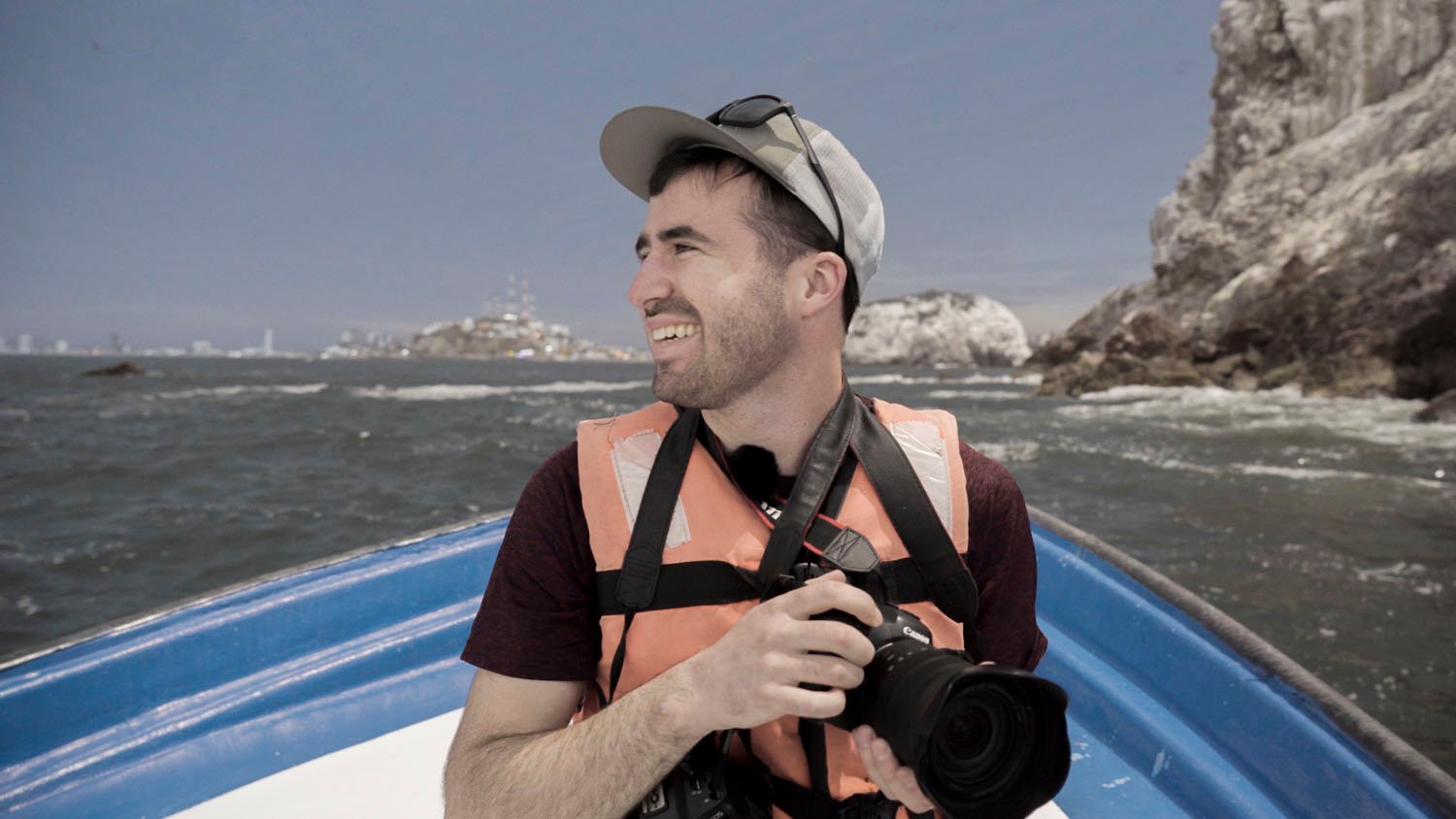 A smiling man wearing a life vest and a cap holds a camera while sitting in a boat, with a rocky shoreline and water in the background.