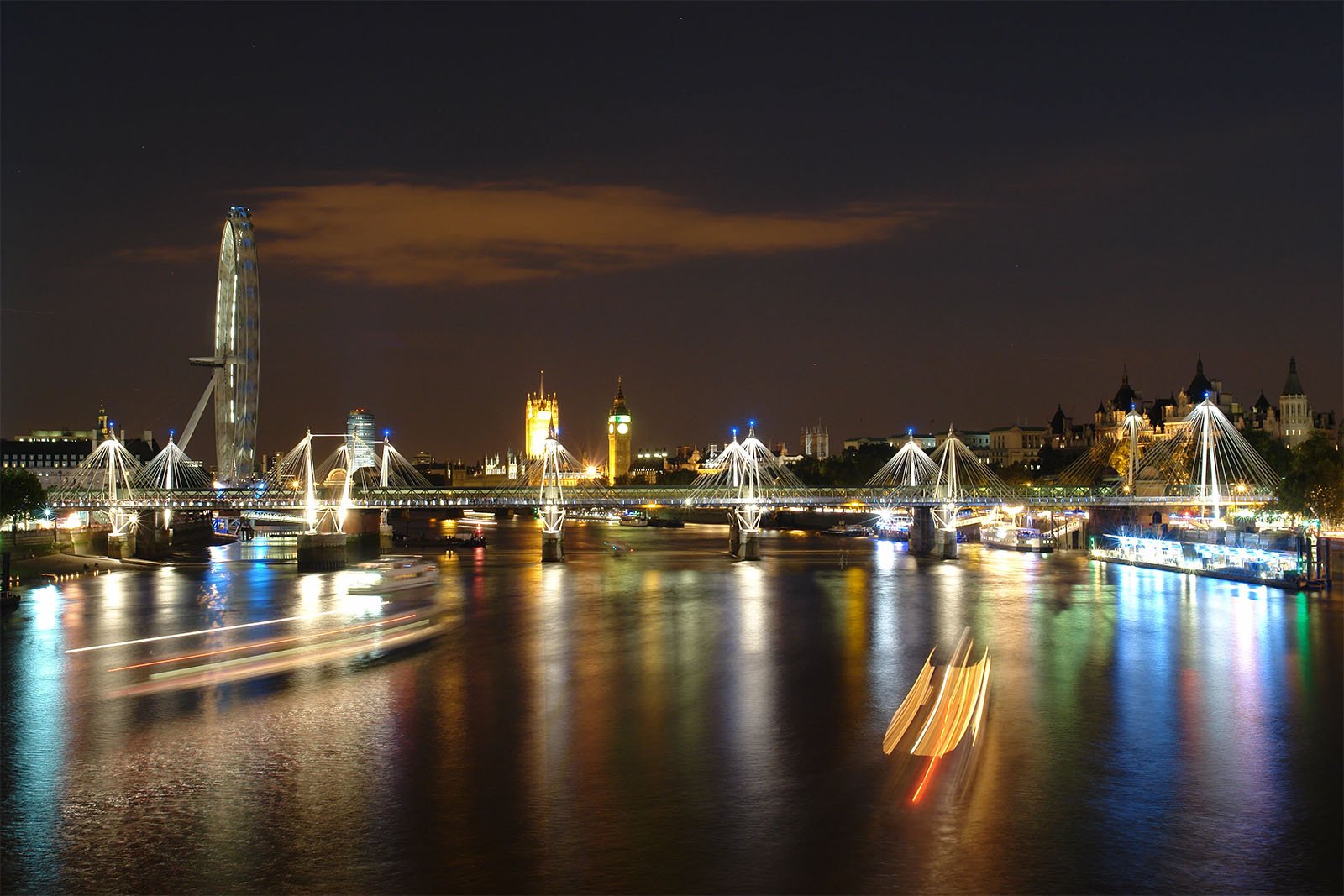 A nighttime view of the river thames in london, featuring the illuminated london eye and houses of parliament, with light trails from moving boats on the water.