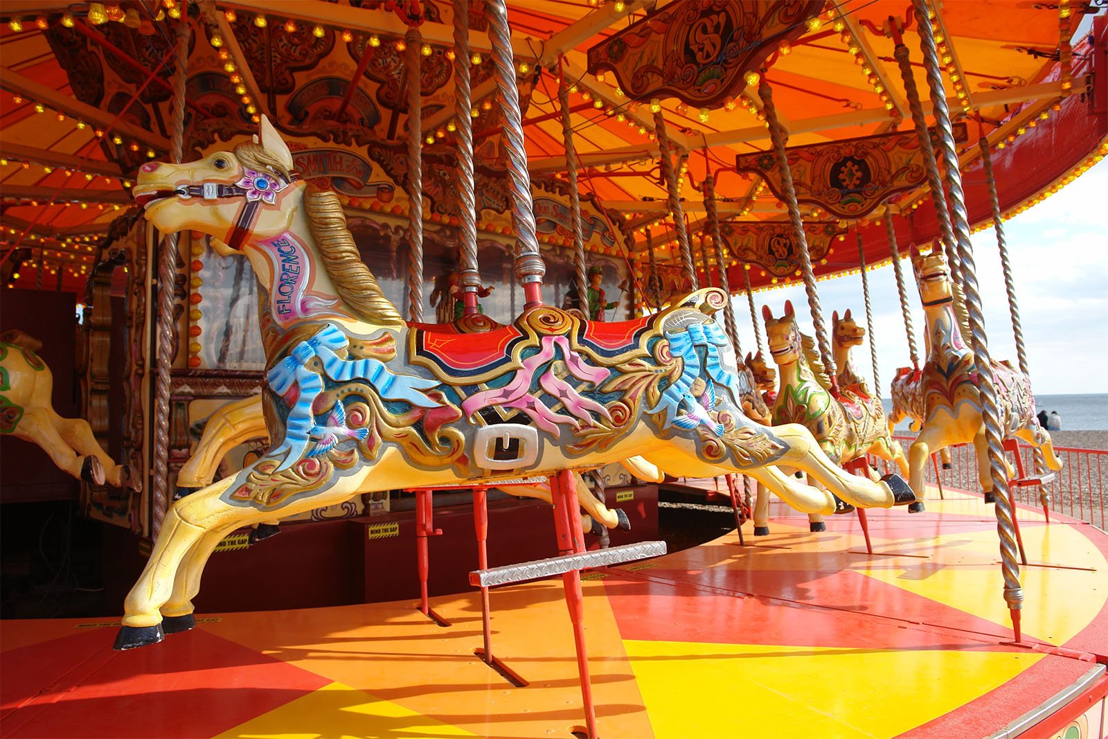 A colorful carousel with elaborate horses on a seaside boardwalk under a cloudy sky. the horses are painted in bright hues with intricate designs and the carousel is framed by red and gold decorations.
